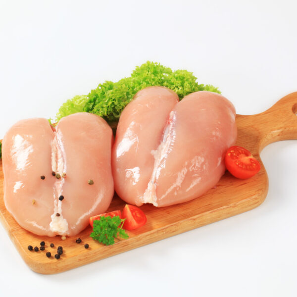 two pieces of raw chicken breast on the wooden cutting board decorated with a leaf of green parsley, lettuce, pepper and red cherry tomatoes
